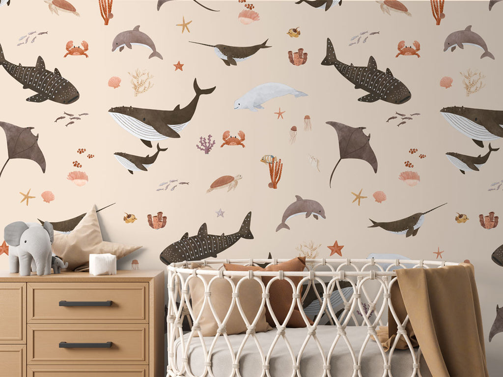 A baby’s nursery with beige Splendid Sea wallpaper, a wicker crib, and marine illustrations of dolphins, whales, and corals.