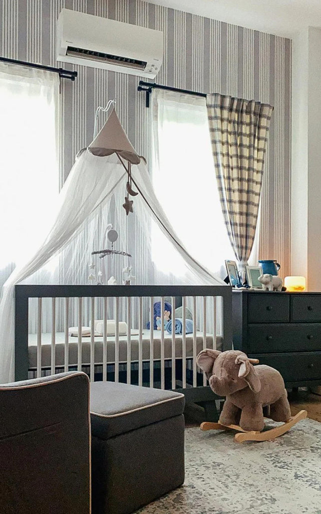 A serene nursery room adorned with Stripes Linen, Pattern Wallpaper in Grey Blue, featuring a white crib with a canopy, a plush elephant toy, an armchair, and a dresser by a window.