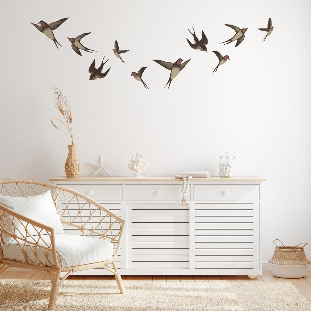 A tranquil room with a white dresser, rattan armchair, and natural woven rug. The wall features Swallows, Wall Decals in a flight pattern, adding an artistic touch. A dried plant vase enhances the calm ambiance.