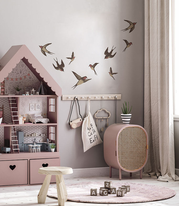 A cozy corner of a room with light grey walls adorned by the Swallows, Wall Decals. The space includes a pink dollhouse-style bookshelf, a round perforated cabinet, and a small wooden stool on a circular pink rug. A plant and draped curtain add warmth.