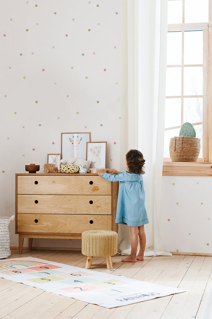 Child’s room bathed in natural light from a large window, highlighting the playful  Tic Tac Toe, Pattern Wallpaper in Multicolor. The cozy ambiance is accentuated by a wooden dresser, creating a simple yet stylish decor.