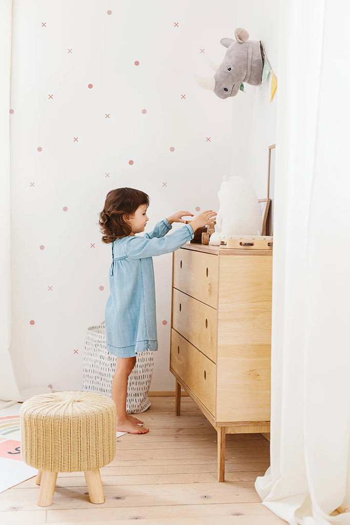 A child reaches for items on a wooden dresser in a light-filled room. The walls, adorned with a pink Tic Tac Toe, Pattern Wallpaper, add a playful touch to the space. A plush toy and woven stool contribute to the room’s cozy and functional decor.