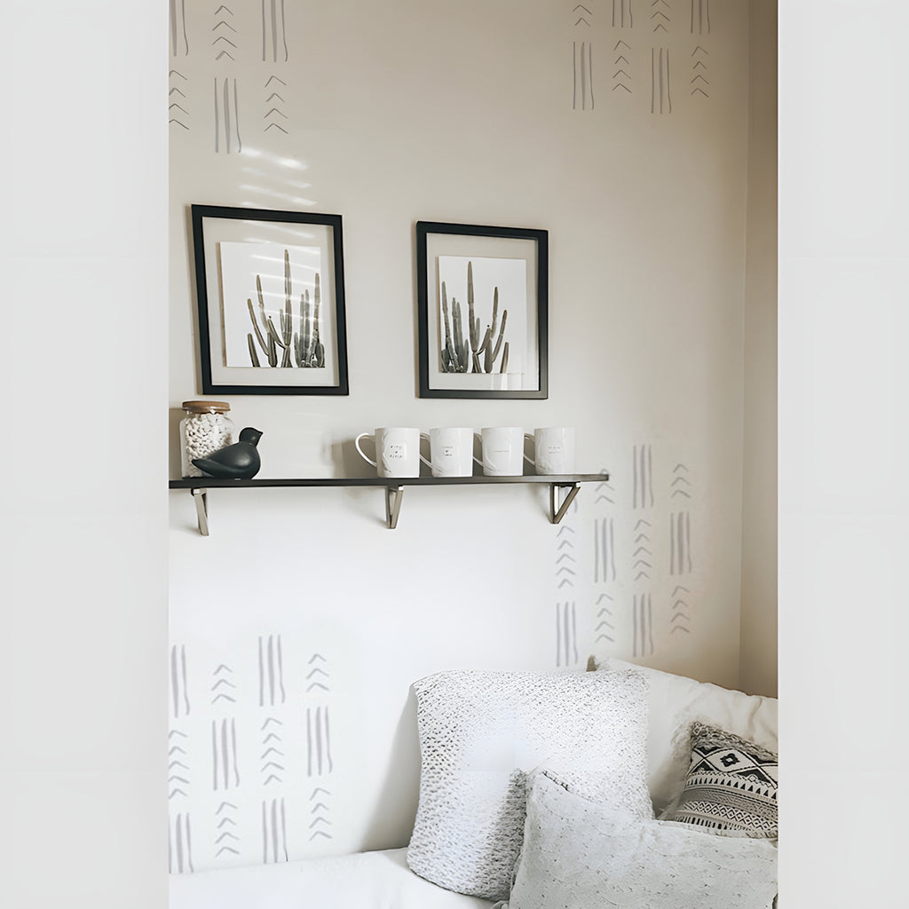 A cozy room corner with a white sofa adorned by a patterned cushion. Above, a floating shelf displays two framed artworks, a decorative bird figurine, and four hanging mugs. The wall features Tribal Geometric Lines, Wall Decals, adding an artistic touch to the space.