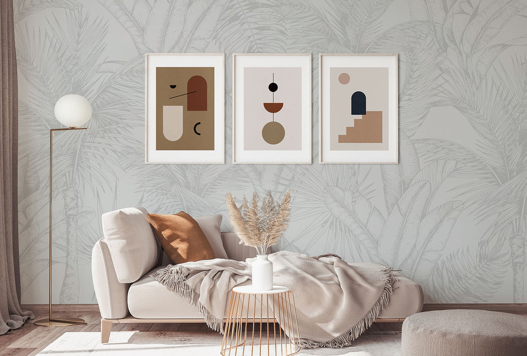 A tranquil living room with Tropics Botanical, Mural Wallpaper in Light Grey. A comfy sofa with a plush throw is under three abstract art frames. A modern lamp and dried pampas grass decor are nearby.