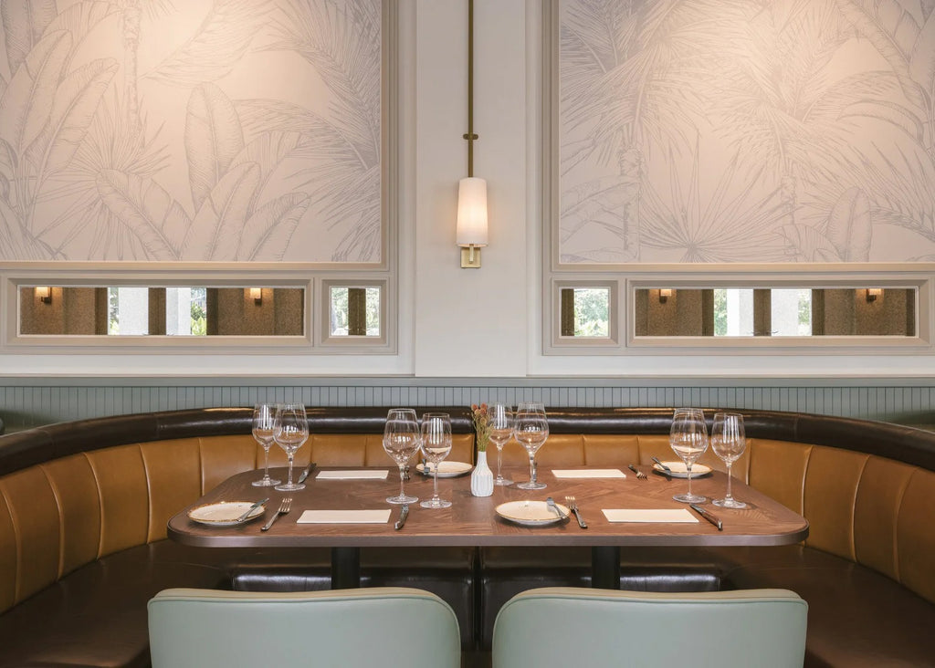 Elegant dining space with Tropics Botanical, Mural Wallpaper in Light Grey, featuring subtle palm leaf patterns. Room includes a curved leather booth, white-clothed tables with glassware, and a chic pendant light.