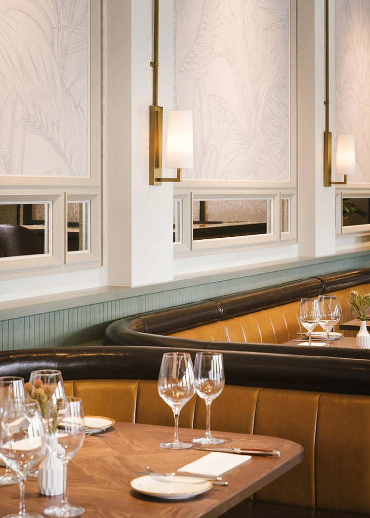 Elegant dining area with tan leather booths, white linen-topped tables set with wine glasses, and a Tropics Botanical, Mural Wallpaper in Grey. Warm lighting from brass sconces adds ambiance.