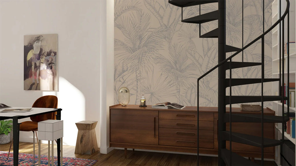 A modern interior room featuring the Tropics Botanical, Mural Wallpaper in Sand with a large leaf pattern. Room includes a wooden desk, spiral staircase, and abstract art.