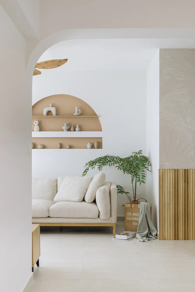 A serene living room corner with Tropics Botanical, Mural Wallpaper in White Nude, featuring a plush white sofa, wooden shelves with decorative items, a potted plant, and a textured wall panel.