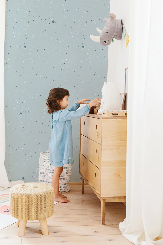 A child reaches for items on a wooden dresser in a light-filled room. The walls, adorned with a Blue Twinkle Twinkle Little Stars, Wallpaper, add a playful touch to the space. A plush toy and woven stool contribute to the room’s cozy and functional decor.