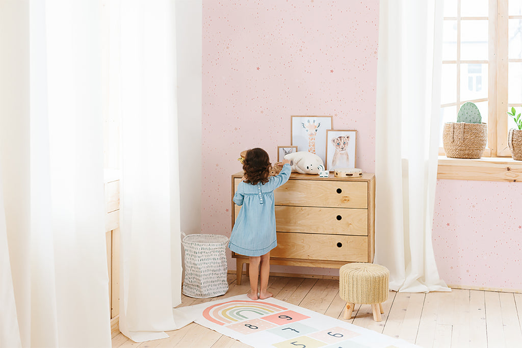 A child in a blue dress is in a bright, minimalist room. The room, adorned with a playful terrazzo-style wallpaper named Twinkle Twinkle Little Stars, Wallpaper in Pink, exudes a serene ambiance. A wooden dresser with animal illustrations, a woven basket, and a colorful rug with numbers add to the room’s charm.