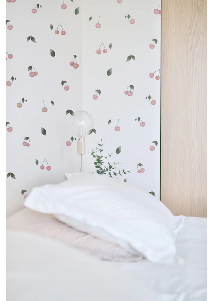 A tranquil bedroom with Watercolour Cherries, Pattern Wallpaper. The room features a cozy bed with a white pillow, a wooden headboard, and a minimalist white lamp. A touch of greenery adds a fresh vibe to the space.