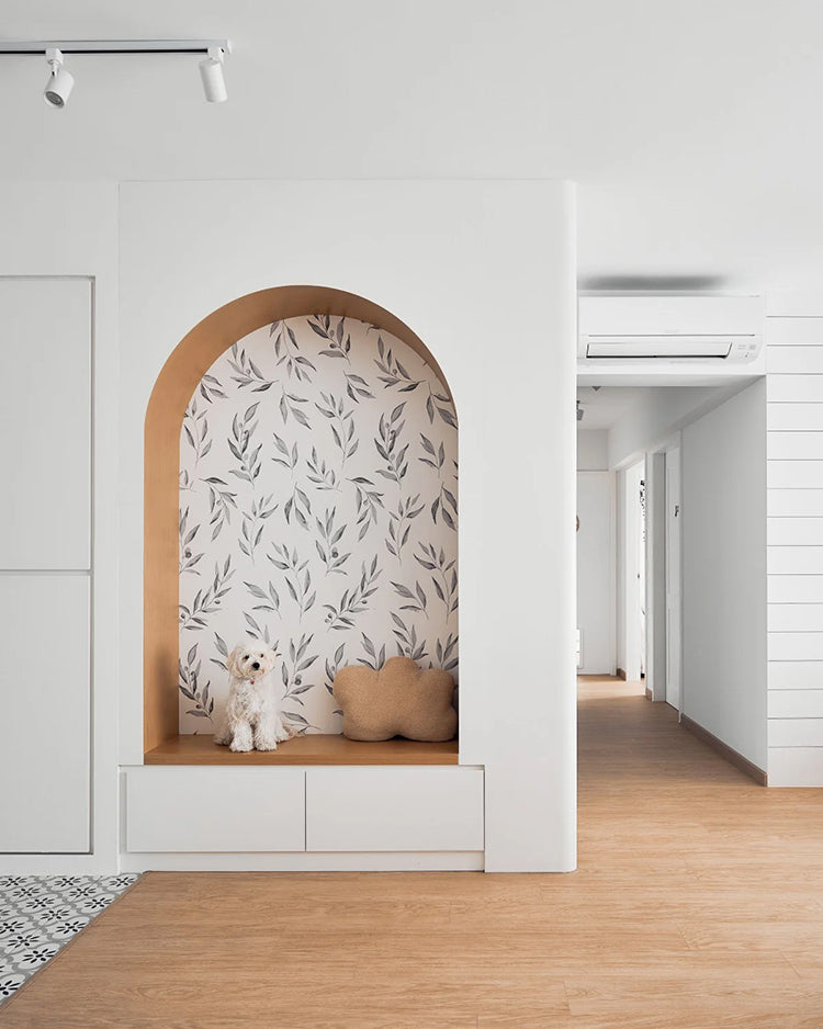 Contemporary room with white walls, wooden flooring, and track lighting. An arched nook with Watercolour Foliage by Olivia, Pattern Wallpaper in Grey pattern wallpaper houses a built-in bench with beige cushions and two fluffy white dogs.
