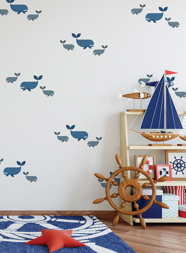 A charming children’s room with a nautical theme. The white walls feature Whales, Wall Decals, creating an oceanic ambiance. A wooden shelf displays a model sailboat and other maritime decor. Adjacent to the shelf, a ship’s wheel leans against the wall. Completing the scene, a star-patterned rug covers the floor, inviting young adventurers to explore the deep blue sea.