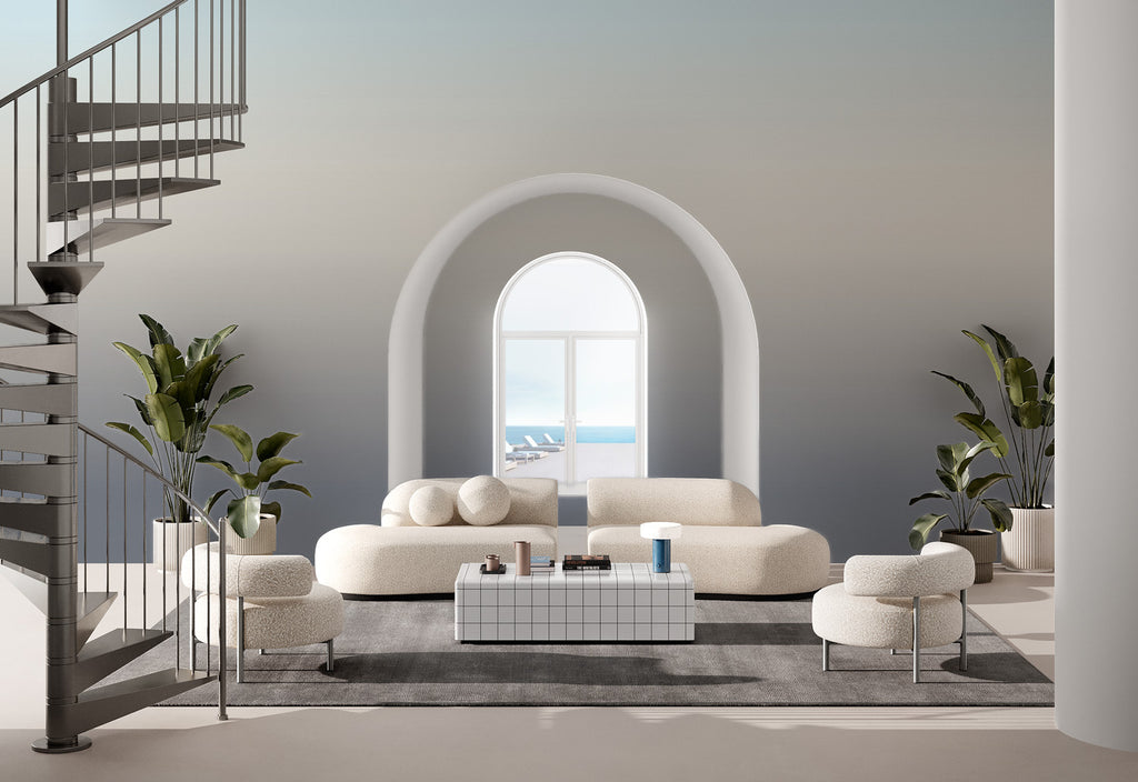 A serene living room with a large arched window revealing a calming seascape. The space features a white, plush modular sofa adorned with round cushions, flanked by potted plants. A spiral staircase adds an architectural touch to the left. The walls showcase the Luminate Gradient, Ombre Mural Wallpaper in Horizon, transitioning colors.