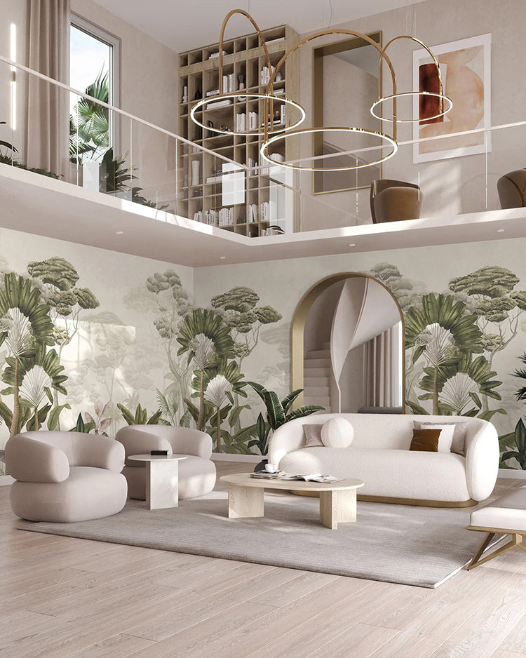 In this spacious, modern living room, natural light floods through tall windows, illuminating a two-level design. The main wall boasts the pale Garden Eden, Tropical Mural Wallpaper in Sand, which transports you to a tropical paradise. The room is furnished with a beige sofa set, a wooden coffee table, and unique circular chandeliers, creating an inviting blend of contemporary aesthetics.