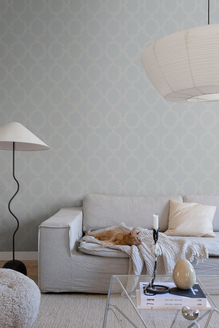 Wilma Wreath Patterned Wallpaper in Dusk Blue Featured on a wall of a living area with a grey sofa with a dirty white fabric and a pillow, along with a glass coffee table, that has a candle and a marble ornament on top, and a lampstand beside it.