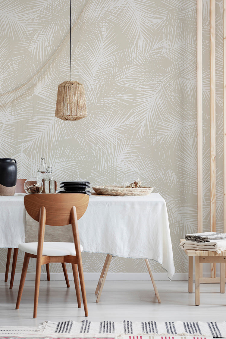 A cozy dining area with a wooden table set, modern decor, and a woven pendant light. The room is illuminated by natural light, highlighting the Lilo Fern, Tropical Pattern Wallpaper in Sand