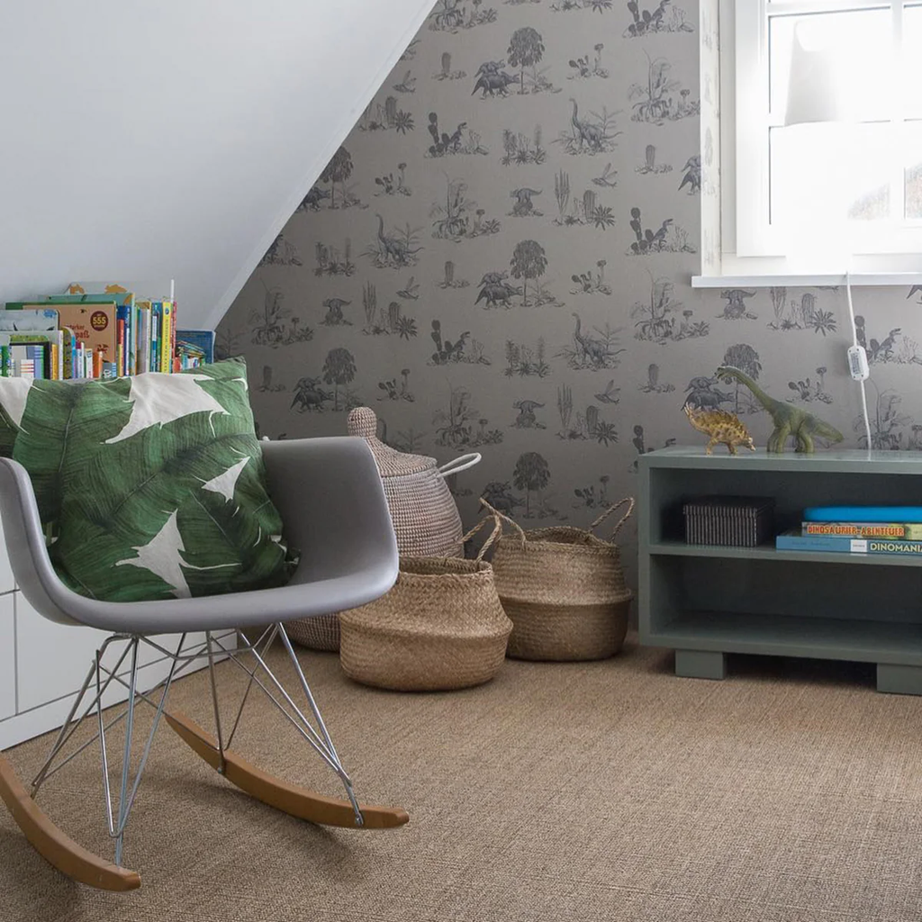 Classic Dino, Pattern Wallpaper in Grey featured on a wall bathed in natural light from a window. A modern gray rocking chair with wooden rockers is adorned with a leaf designed cushion. A small dark green bookshelf holds books and an ornamental item, and woven baskets add a homely touch.