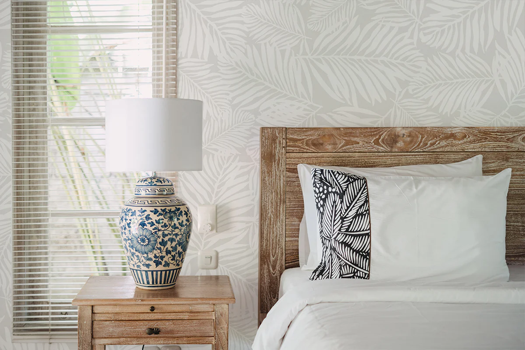 Elegant bedroom with Heather Fern, Pattern Wallpaper in Light Grey, featuring white and beige botanical design. Includes a wooden bed, white bedding, patterned pillow, and a classic blue and white porcelain lamp on a wooden nightstand.
