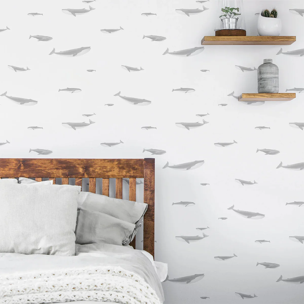 Serene bedroom scene with a wall adorned by ‘Ocean Whale, Pattern Wallpaper’, featuring grey whale silhouettes on a white backdrop. A wooden headboard, cozy bedding, and two wooden shelves with decor items are visible