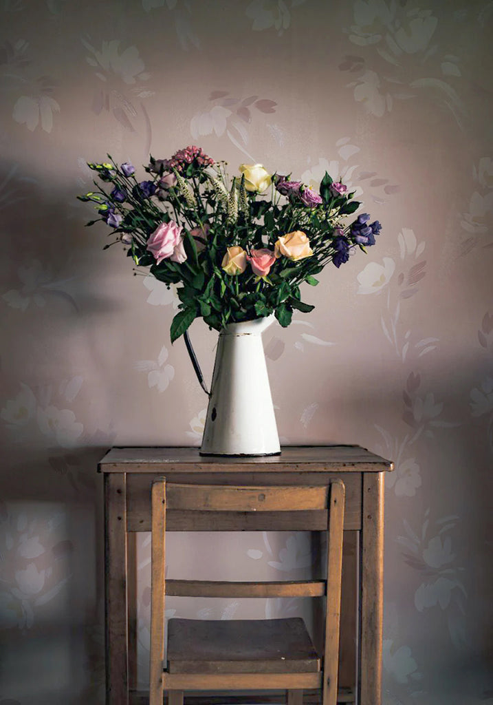 Rebecca, Floral Pattern Wallpaper in Nude featured on a wall of a room with wooden chair and table, on top of it is a vase with a bouquet of flowers