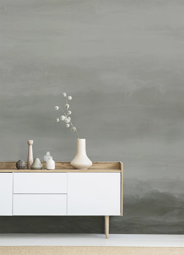 In a room, a wall features the Vista Watercolor Ombre Wallpaper in Stratos Grey, creating a striking contrast with a white table. The table is adorned with multiple ceramic vases, adding to the room’s aesthetic appeal.
