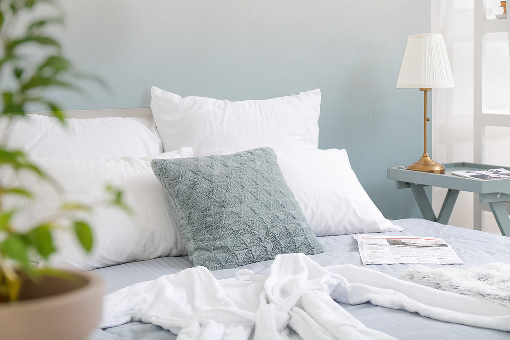 A serene bedroom featuring Tessa, Ombre Wallpaper. The room is highlighted by a neatly made bed with white and patterned pillows, a lamp on a bedside table, and a newspaper adding a personal touch.