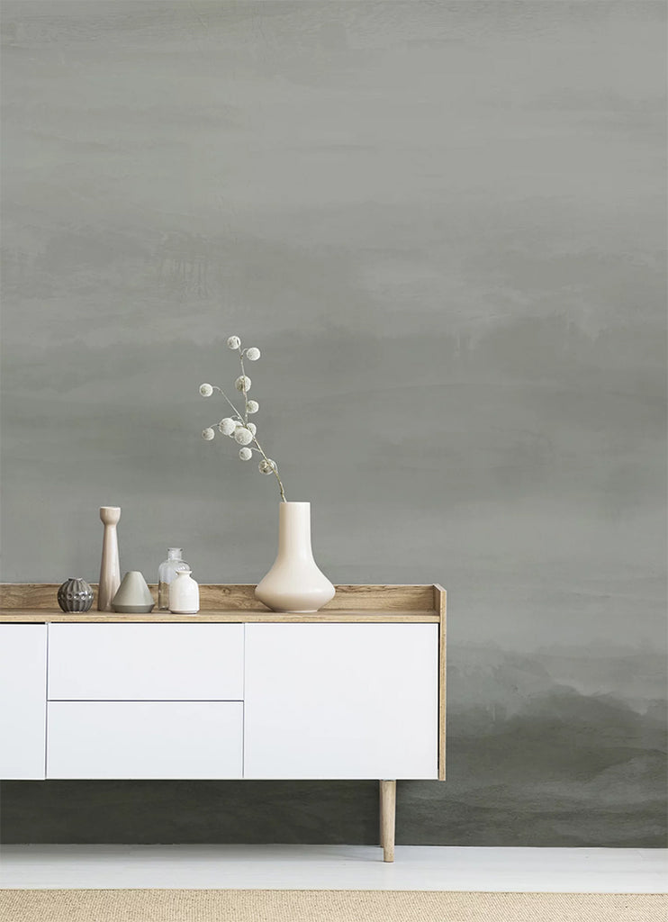 In a room, a wall features the Vista Watercolor Ombre Wallpaper in Stratos Grey, creating a striking contrast with a white table. The table is adorned with multiple ceramic vases, adding to the room’s aesthetic appeal.