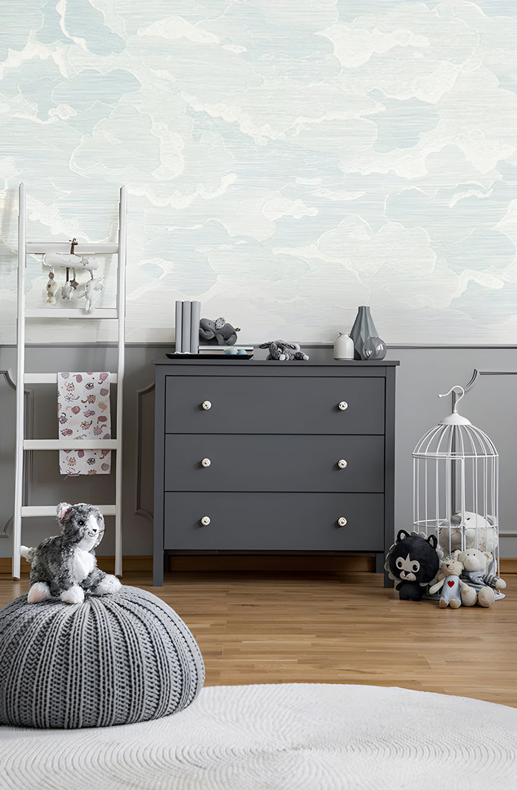 Illustrated Clouds, Mural Wallpaper in Blue, featured on a room wall, that brings whimsical and serene atmosphere . This room is adorned with a grey cabinet, on top of which ceramics are displayed. Plush toys are scattered around the room, adding a playful touch to the space. The blue mural wallpaper with its illustrated clouds brings a whimsical and serene atmosphere to the room.