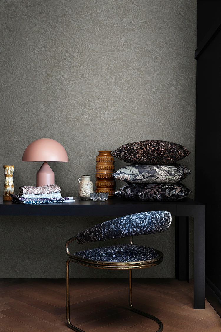 Sea Foam, Wallpaper in Light Grey featured on a wall of a room, with black table that has multiple patterned pillow, and pink lamp, with a patterned chair next to it.