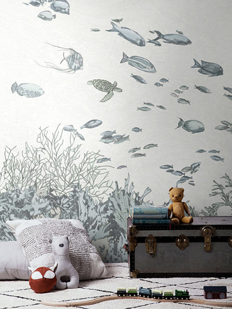Under the Sea, Wallpaper in dusk blue sets the theme in a room that features a grey treasure chest with a brown stuffed toy, and other stuffed toys are comfortably nestled on the pillow. Train toys can be seen scattered on the floor, adding to the room’s playful charm.